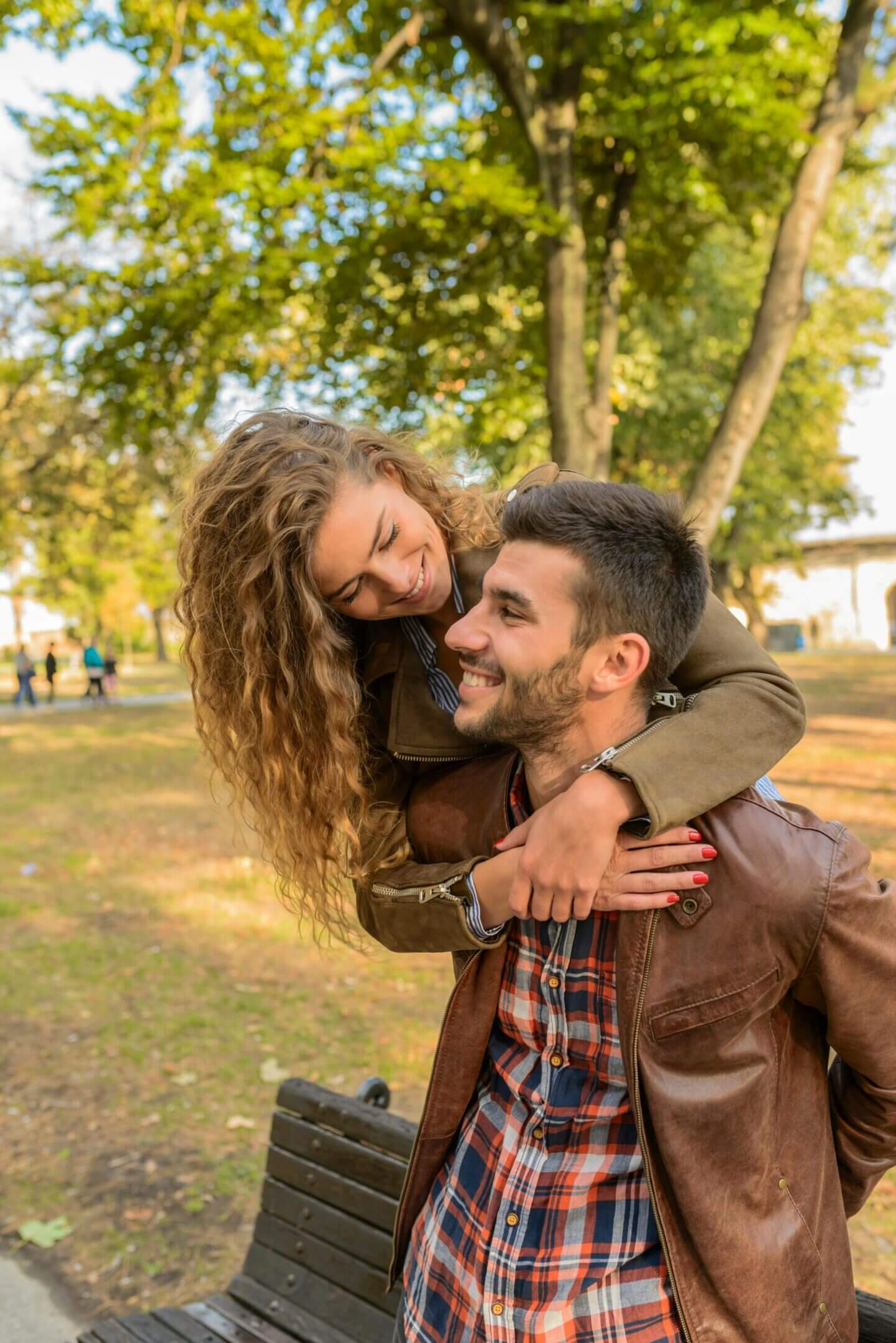 A woman hugs a smiling man from behind in a park.